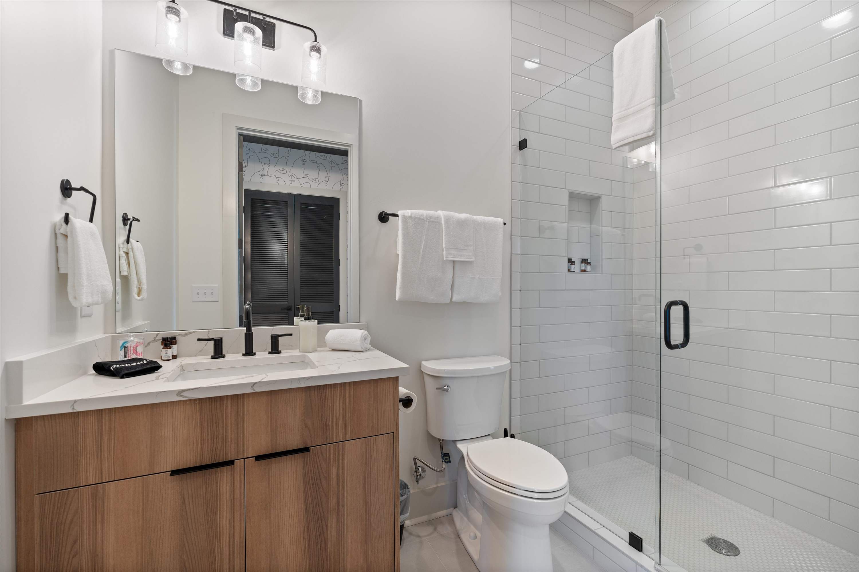 Bright modern bathroom with a light wood vanity, white countertop, and a large mirror with contemporary lighting. A glass-enclosed shower with white subway tiles and a white toilet complete the space.