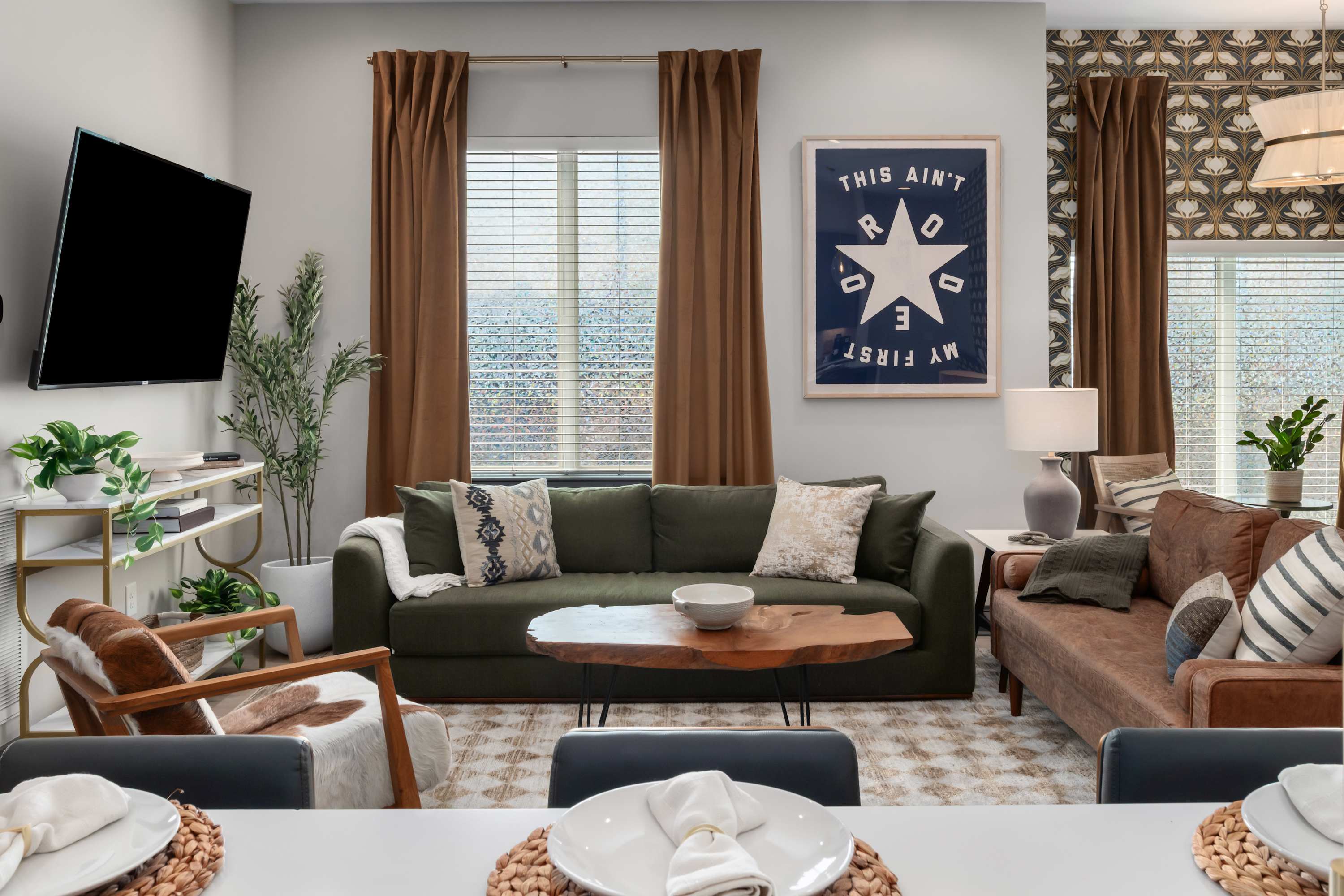 A cozy living room features a dark green sofa with patterned pillows, a live-edge wood coffee table, and a brown leather sofa. A large framed art print hangs above the green sofa, and brown curtains frame windows with blinds.