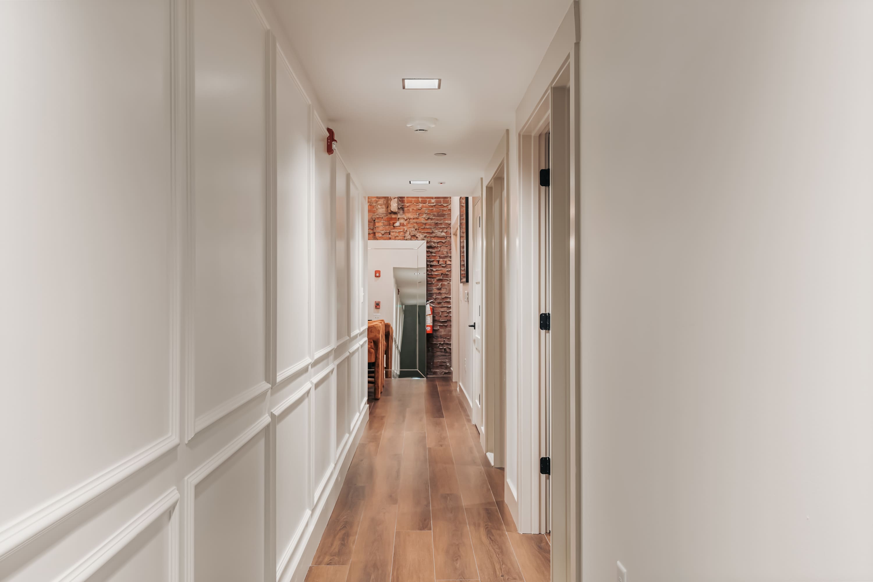 A long, well-lit hallway with white wainscoting on the walls and dark wood-look flooring. Multiple white doors line the hallway, leading to various rooms, with a glimpse of exposed brick at the far end.