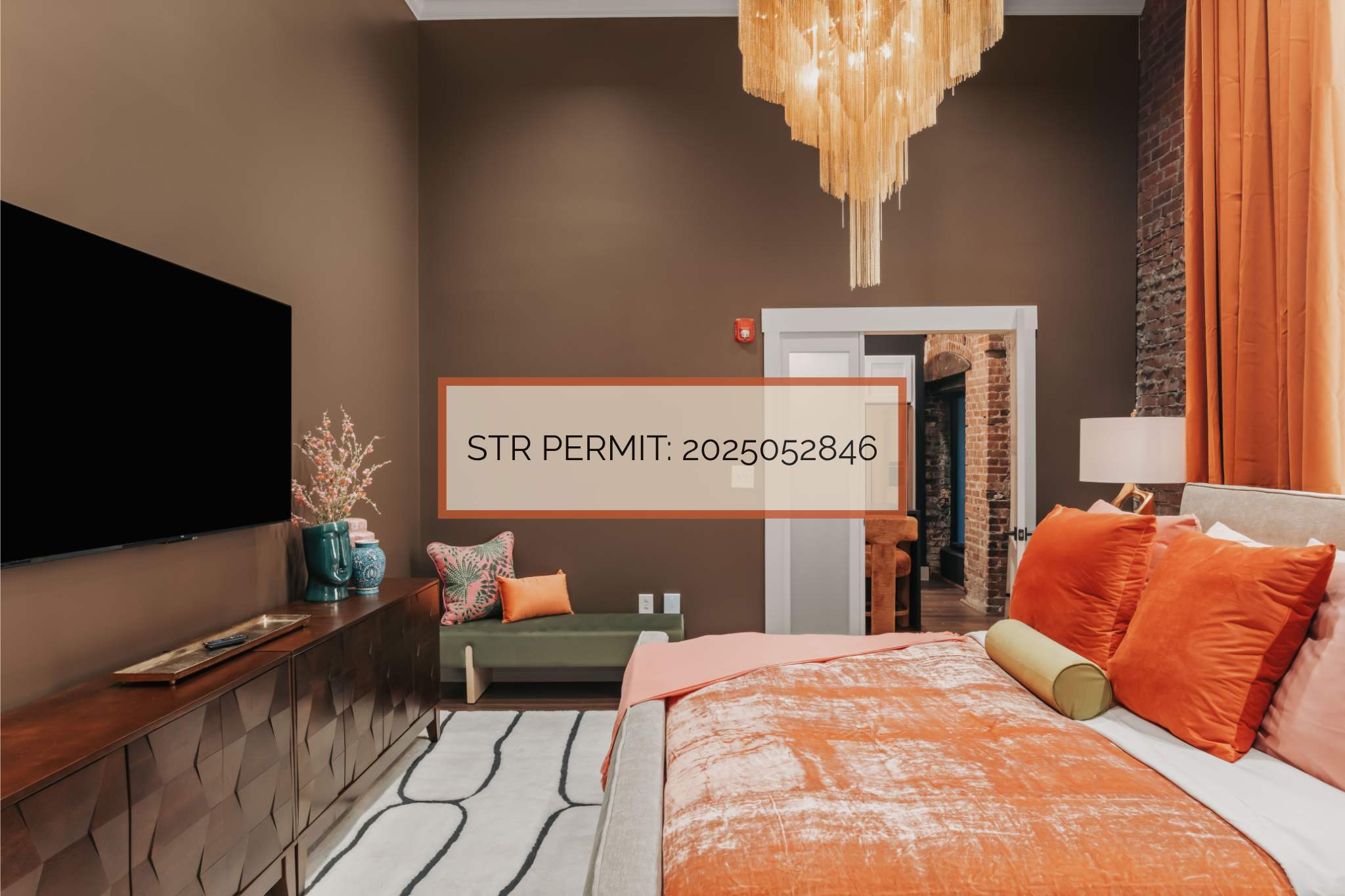 Cozy bedroom with a bed featuring orange and pink bedding, a dark brown accent wall, a large flat-screen TV on a wooden console, and a unique fringed chandelier. A permit number is overlaid on the image.