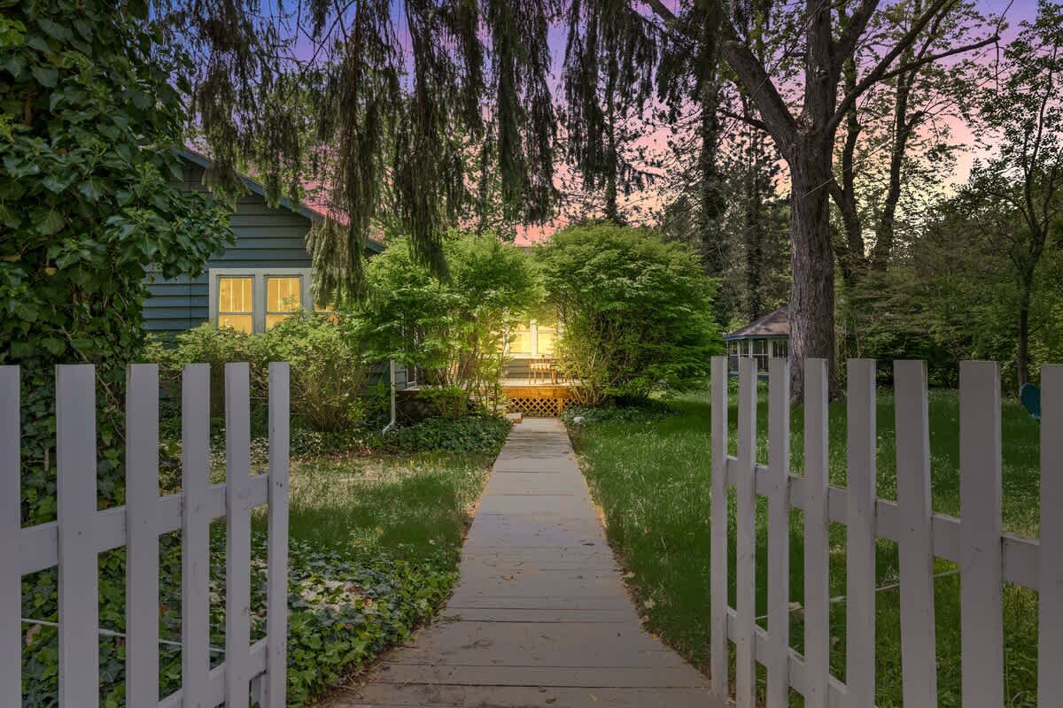 A white picket fence gate opens to a wooden walkway leading to the illuminated blue house, nestled among trees and bushes under a twilight sky with hints of pink and purple.