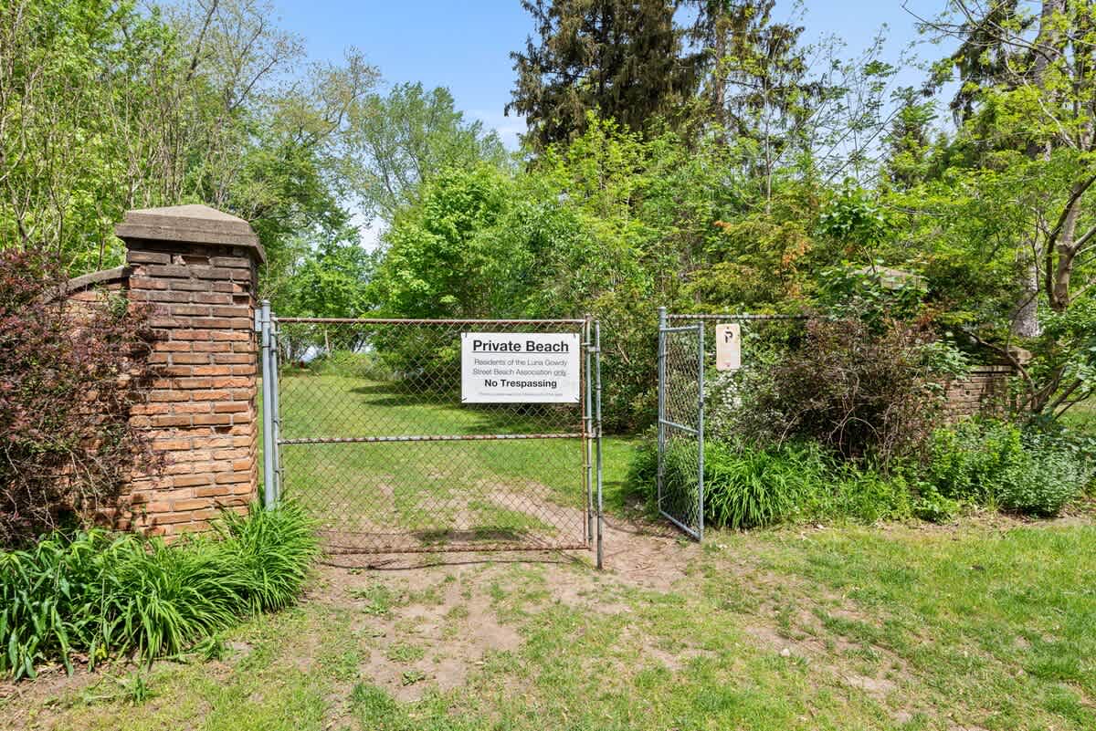A chain-link fence gate with a "Private Beach" sign is set between brick pillars, indicating access to a beach area, surrounded by green foliage and trees.