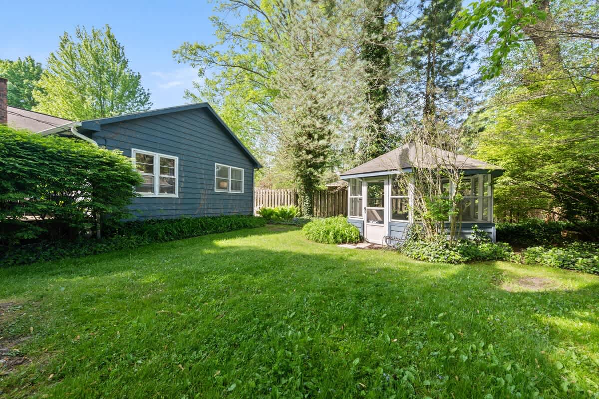 A lush green lawn stretches between a blue-sided house with white trim and a charming screened gazebo, surrounded by mature trees and dense foliage under a bright sky.