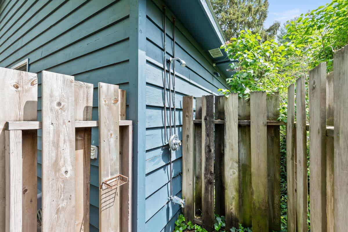 Outdoor shower setup against the blue siding of the house, partially enclosed by a rustic wooden fence. The shower fixture and a small soap dish are visible, surrounded by lush greenery.