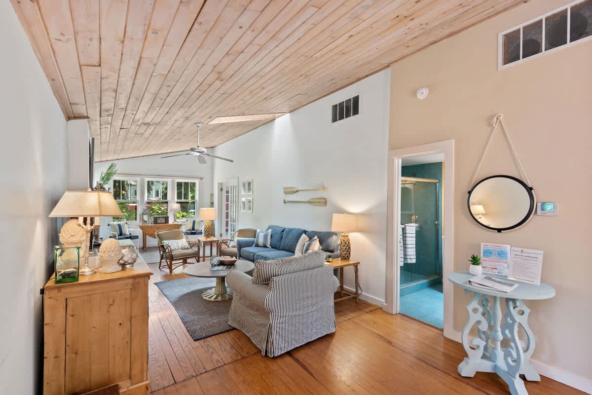 A spacious living room with light wood plank ceilings and hardwood floors features a blue sofa, a striped armchair, and a round coffee table, with a glimpse of a bathroom entrance.