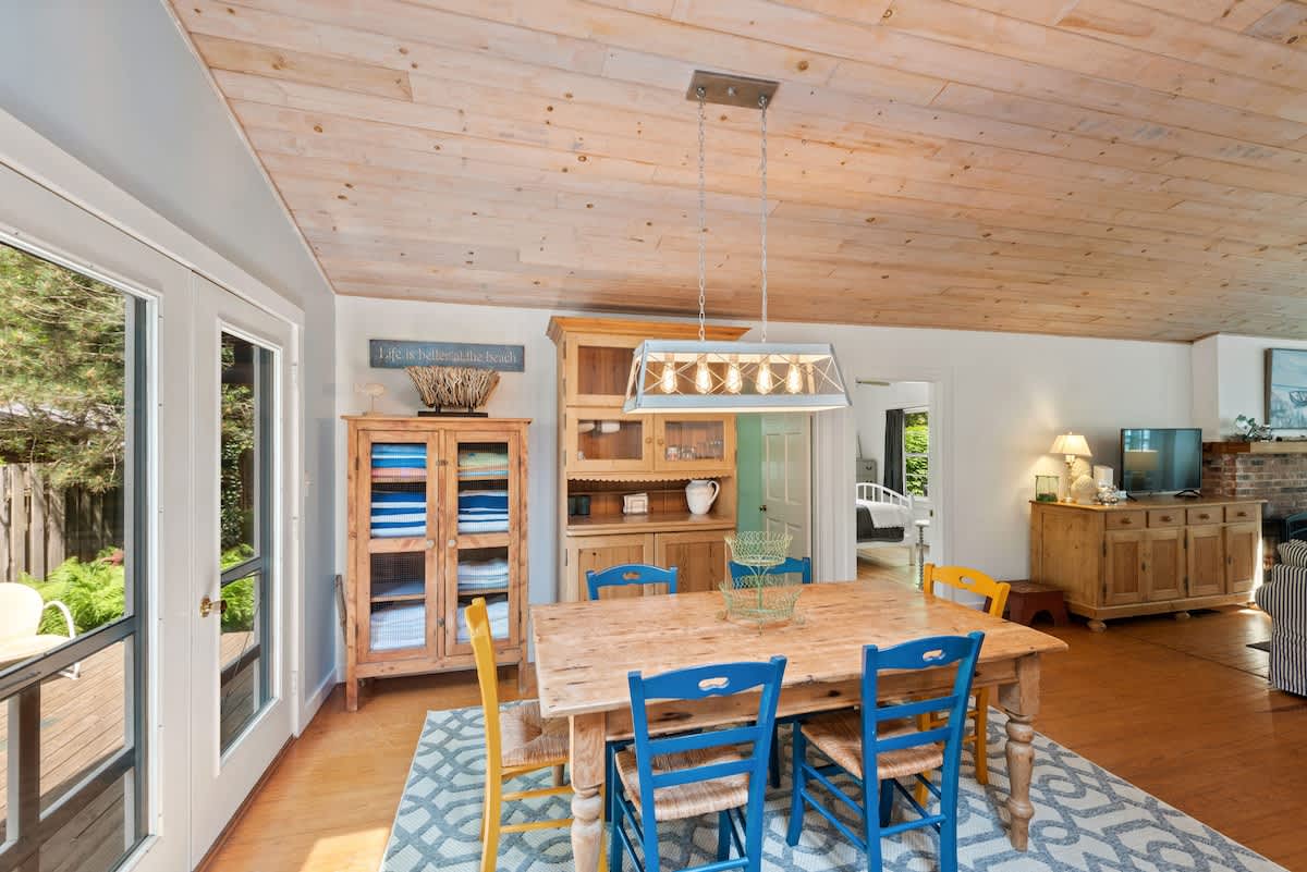 A rustic dining area features a large wooden table with blue and yellow chairs, a light wood plank ceiling, and a geometric rug, with French doors opening to an outdoor deck.