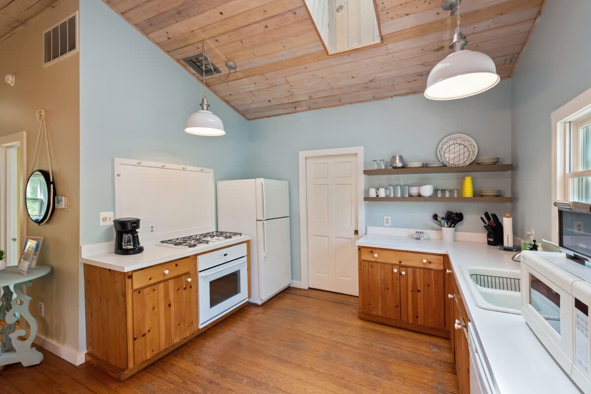 Another angle of the kitchen, highlighting the white gas range with oven, white refrigerator, and coffee maker on white countertops. Light wood cabinets and open shelves are set against light blue walls and a wood-paneled ceiling.