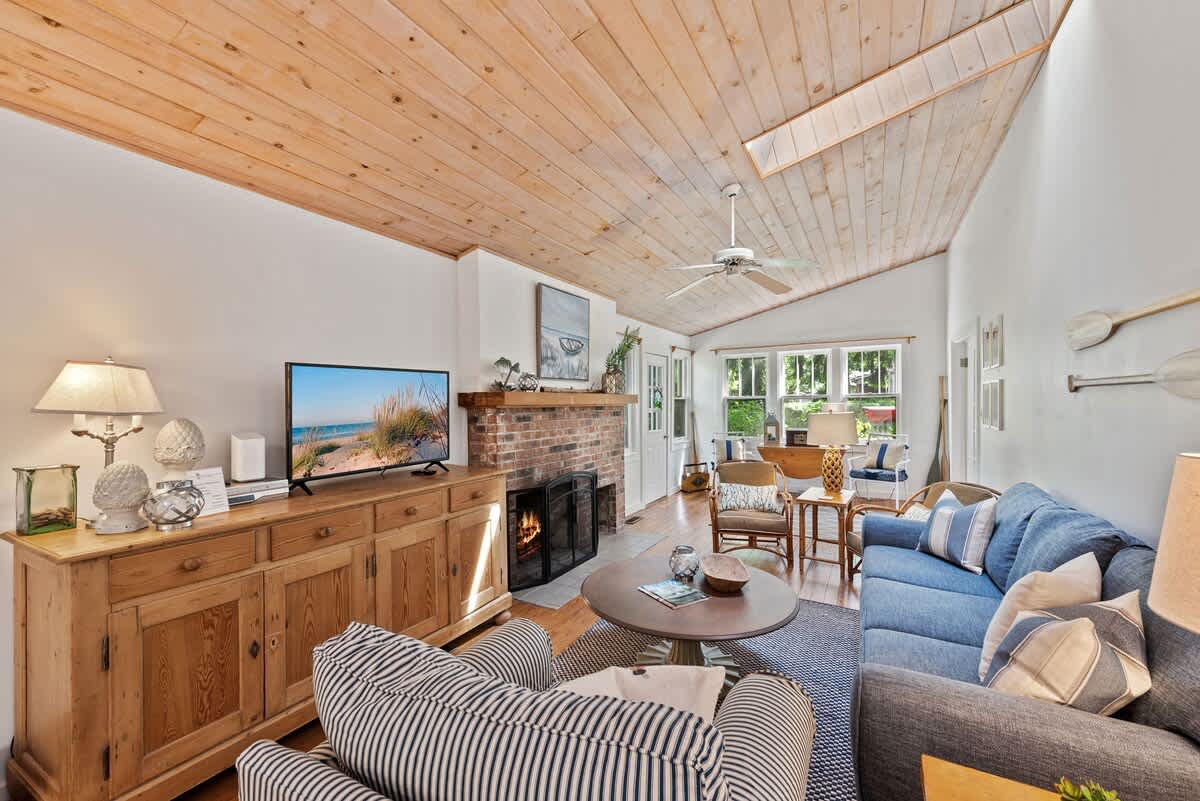 A cozy living room features a blue sofa, a striped armchair, a wooden console table with a TV, and a brick fireplace, all under a light wood plank ceiling with a skylight.