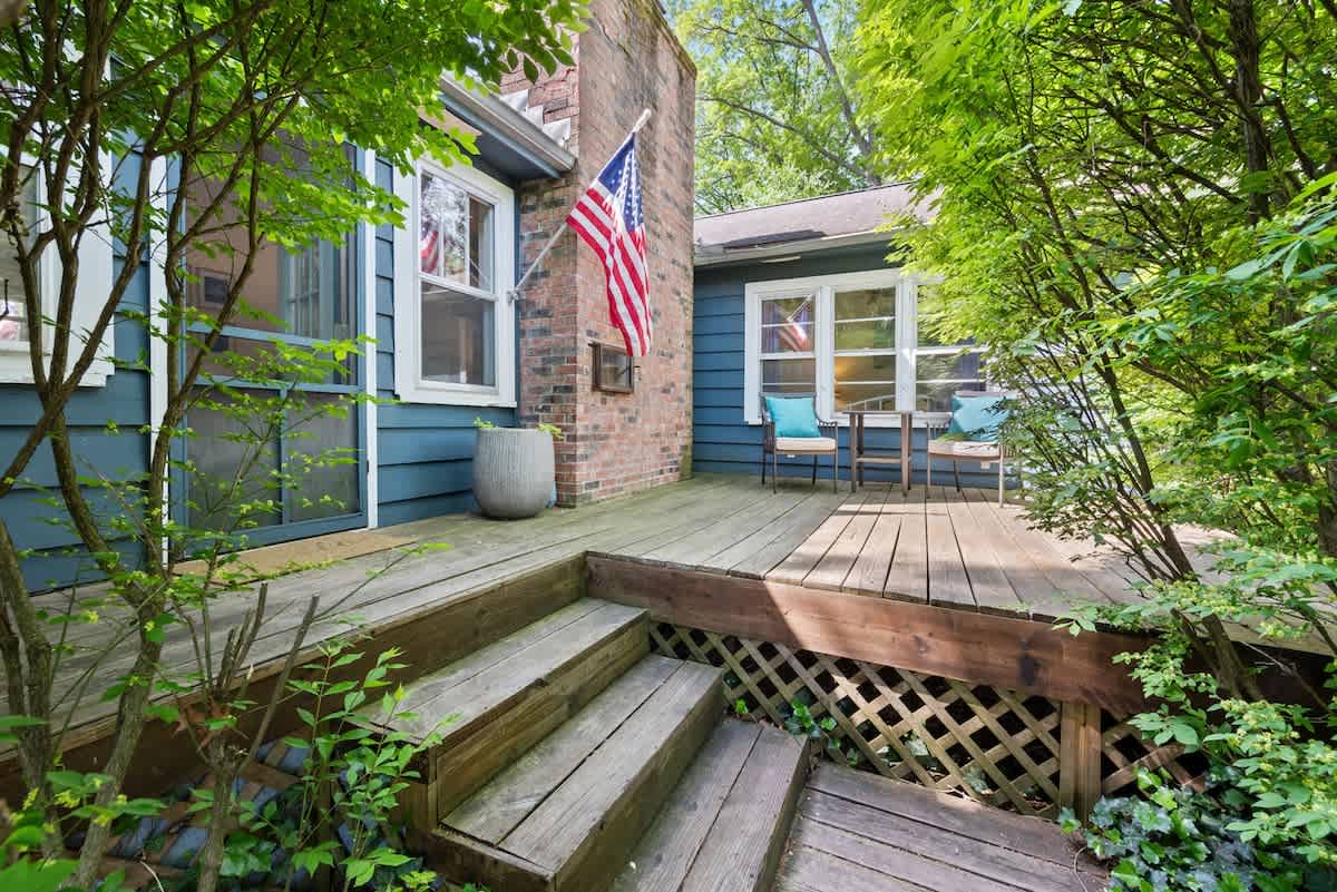A rustic wooden deck with steps leads up to a blue-sided house with a brick chimney, adorned with an American flag, and surrounded by dense green foliage.