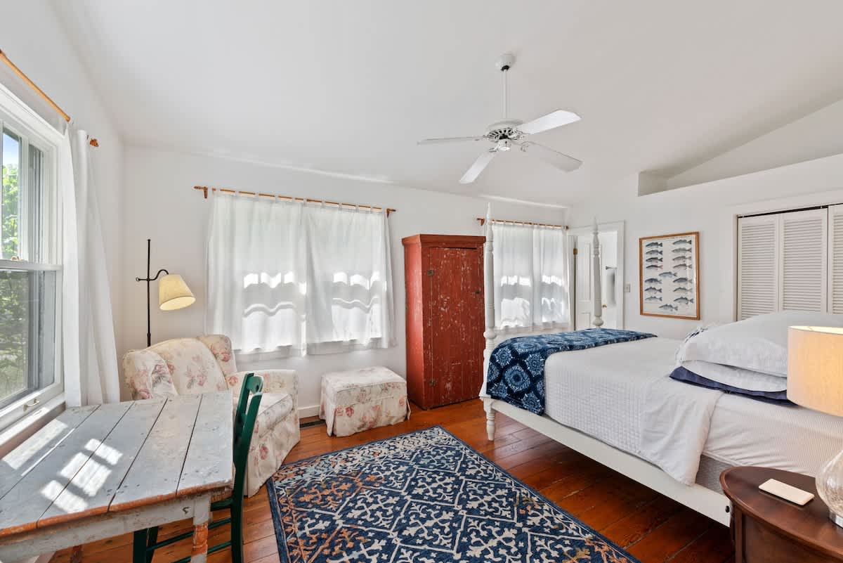 Serene bedroom with white walls and dark wood floors, featuring a white queen bed with blue and white bedding. A vintage red cabinet, a small wooden table, and a ceiling fan add character to the space.