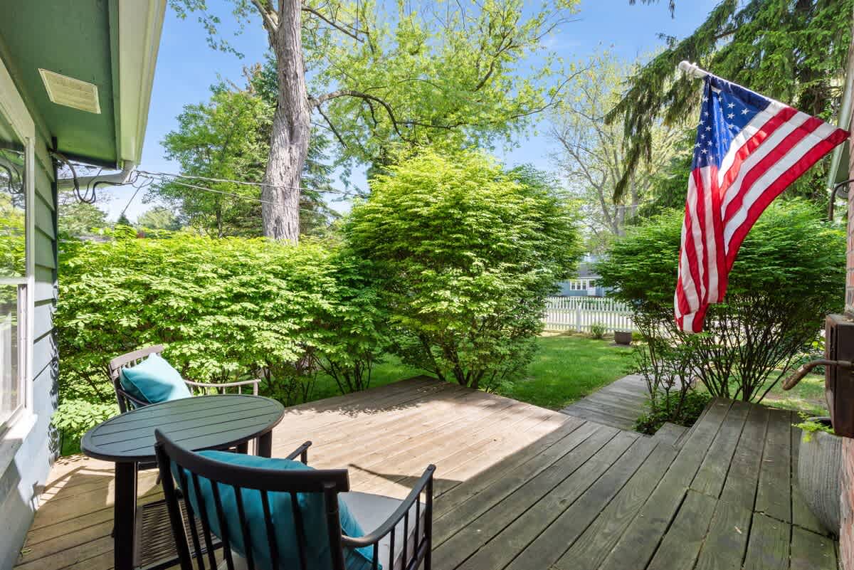 A small wooden deck area with a black metal bistro set and teal cushions is surrounded by lush green bushes and trees, with an American flag hanging from the house.