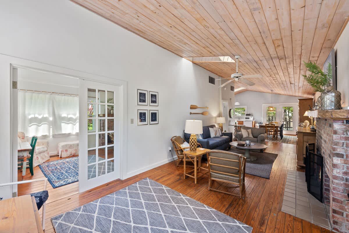 An open-concept living space with light wood plank ceilings and hardwood floors connects to a sunroom through French doors, featuring a blue sofa and a brick fireplace.