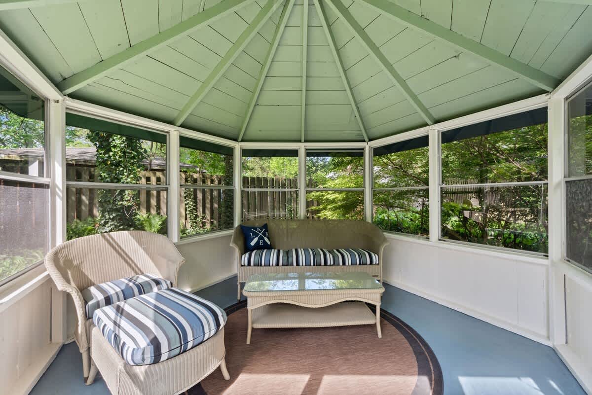 The interior of a screened gazebo features a light green vaulted ceiling, a wicker sofa with striped cushions, a matching armchair, and a glass-top coffee table on a round rug.