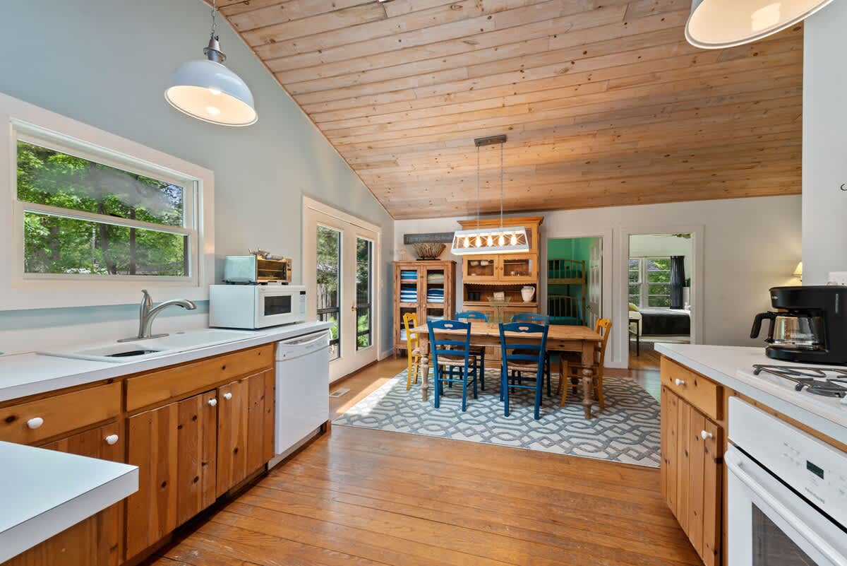 Inviting dining area featuring a large wooden table with blue chairs on a patterned rug, illuminated by a unique chandelier. Double doors lead to the outdoors, complementing the light blue walls and vaulted wood-paneled ceiling.