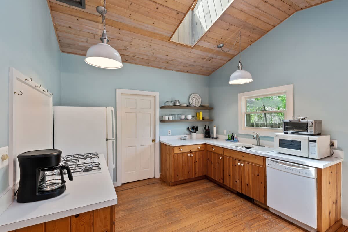 Detailed view of the kitchen with light wood base cabinets, white countertops, and white appliances including a refrigerator, stove, and dishwasher. Open shelving displays dishes, all under a vaulted wood-paneled ceiling and light blue walls.