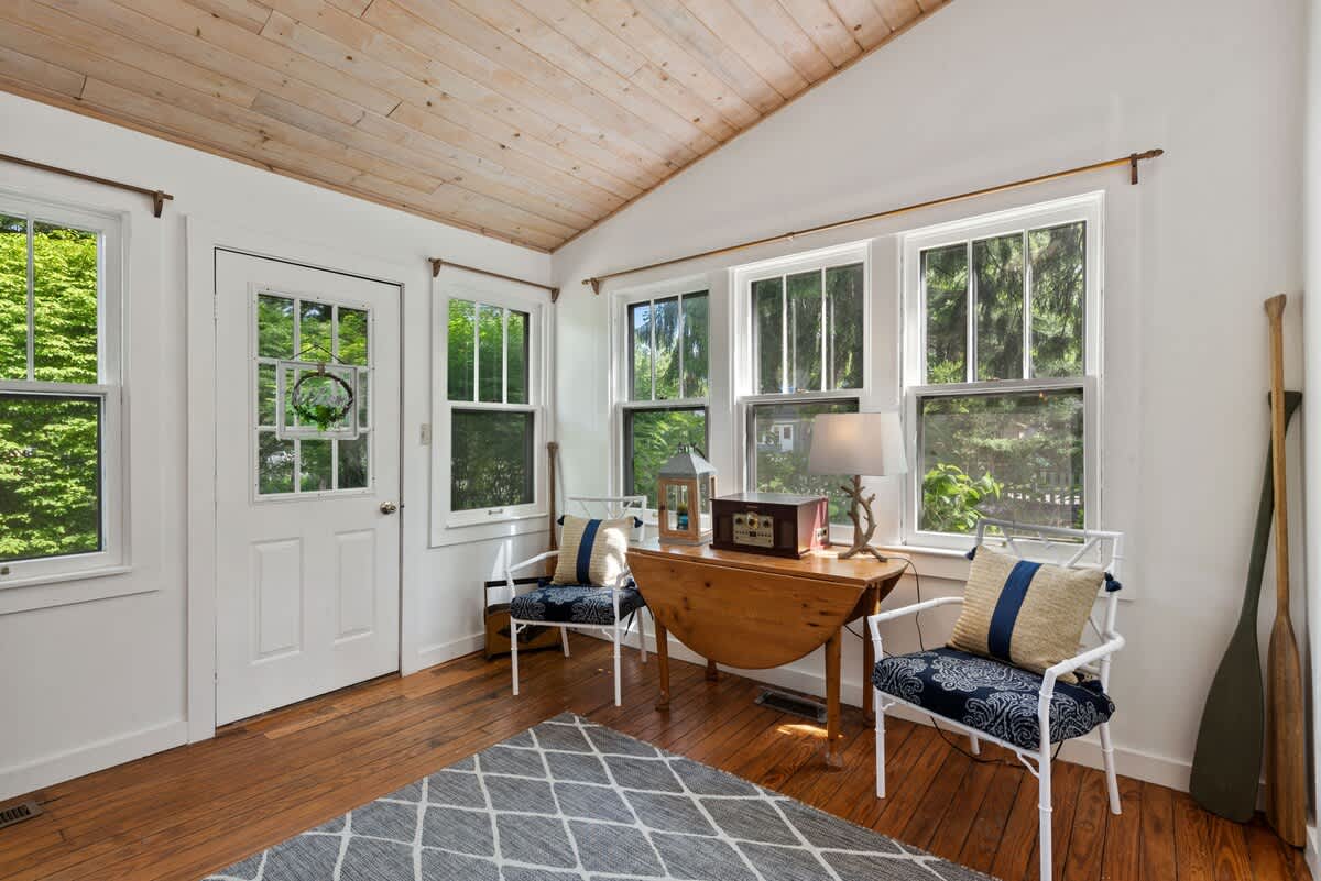 A bright sitting area features light wood plank ceilings, hardwood floors, two white chairs with blue patterned cushions, and multiple windows overlooking lush greenery.