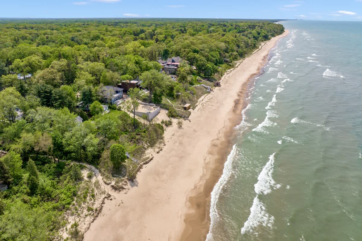 An aerial view captures a wide sandy beach bordering a vast blue lake with gentle waves, backed by a dense green forest and scattered houses along the coastline.
