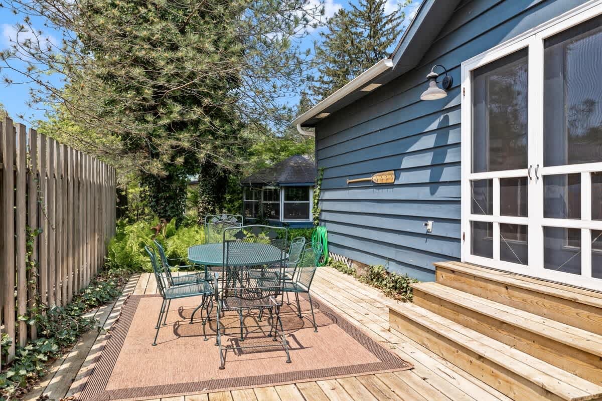 Inviting outdoor wooden deck furnished with a metal patio table and chairs, placed on a brown outdoor rug. The deck is bordered by a wooden fence and lush trees, with a screened door leading into the house.