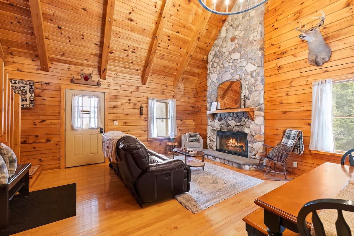 An expansive view of the rustic living room, showcasing the large stone fireplace, vaulted wood-paneled ceilings, and a brown leather sofa. The open-concept space connects to an entry area on the left.