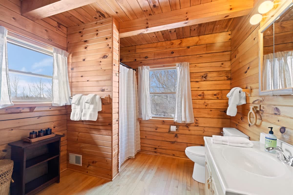 A bright bathroom with light wood plank walls and light wood floors, featuring a white vanity with a single sink and a toilet. Two windows with white curtains provide natural light, and a shower area is visible with a white curtain.