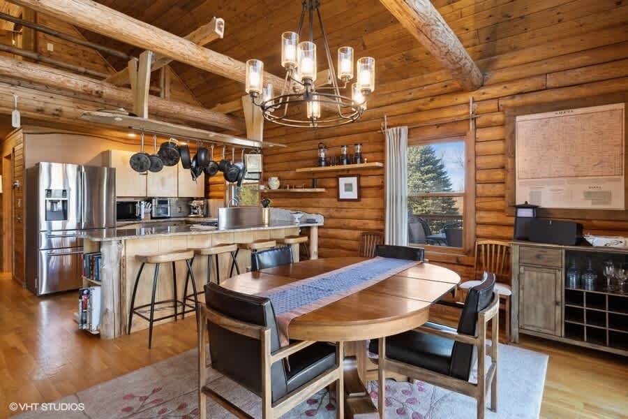 A spacious open-plan dining area in a log cabin features a large wooden oval table with chairs, a modern chandelier, and large windows, with the rustic kitchen island and appliances visible in the background.
