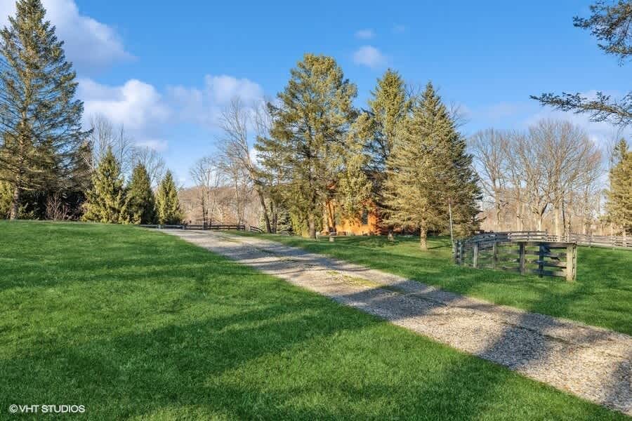 Long gravel driveway winding through a well-maintained green lawn, flanked by tall evergreen trees and a rustic wooden fence, leading towards the house in the distance.