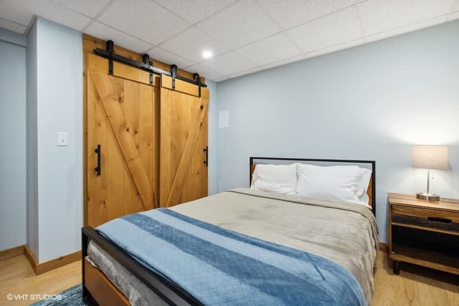 A cozy bedroom features a metal-frame bed with a blue and beige comforter, a wooden nightstand with a lamp, and light blue walls. Large wooden barn doors with black hardware serve as a prominent feature, adding a rustic touch.