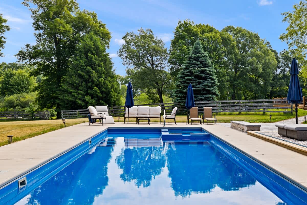 A large rectangular outdoor swimming pool with bright blue water is surrounded by a concrete patio, furnished with various lounge chairs and umbrellas. The pool area is set within a spacious grassy yard, bordered by mature trees under a clear blue sky.