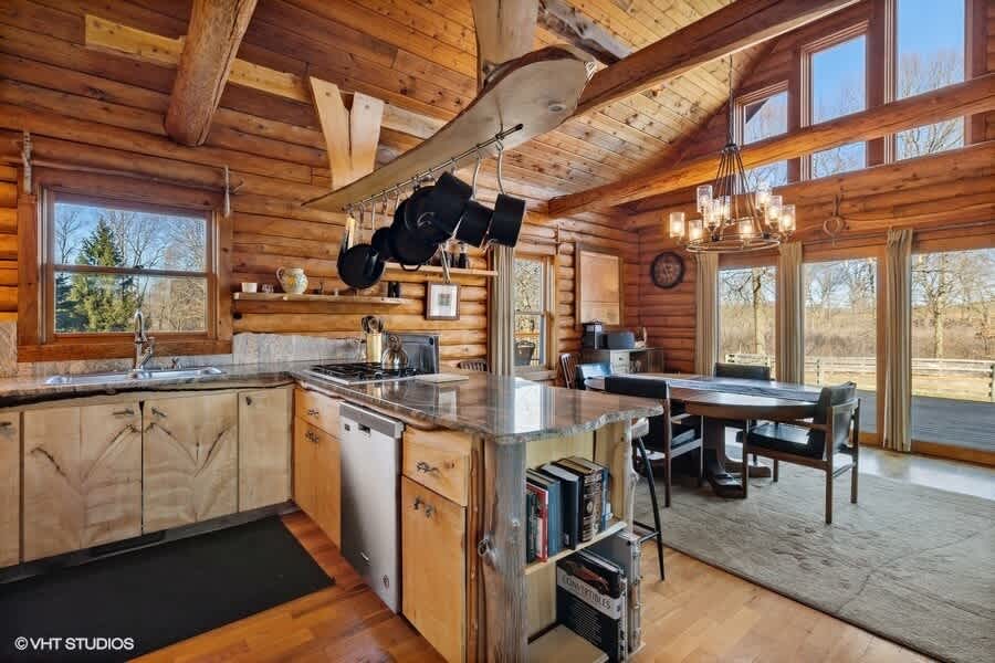 This open-concept log cabin kitchen and dining area showcases a granite-topped island with bar stools, stainless steel appliances, and a rustic pot rack, flowing into the dining space with a wooden table and chandelier.