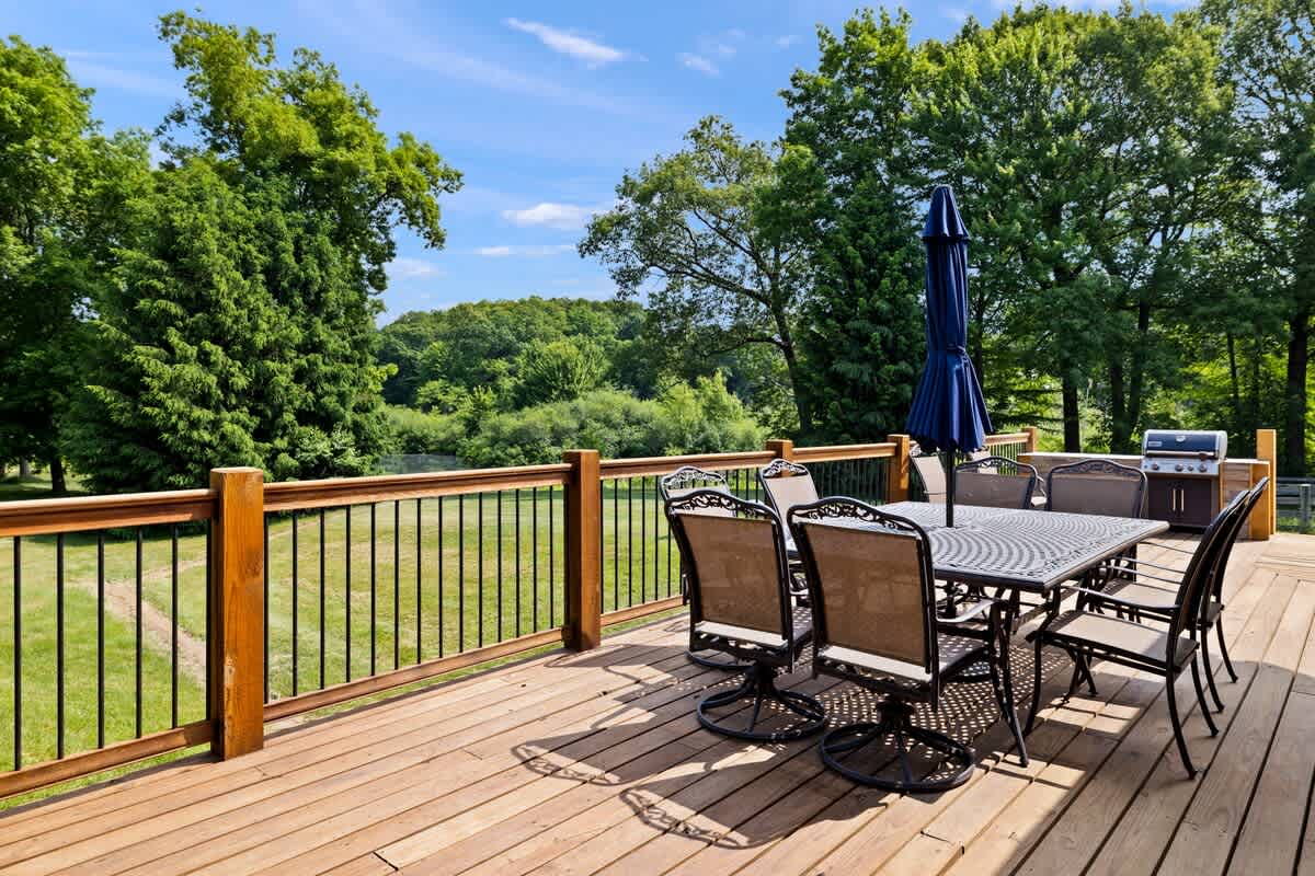 A spacious wooden deck offers an outdoor dining area with a large rectangular table and eight chairs, shaded by a blue umbrella. The deck overlooks a lush green landscape with mature trees and a distant fence under a clear blue sky.