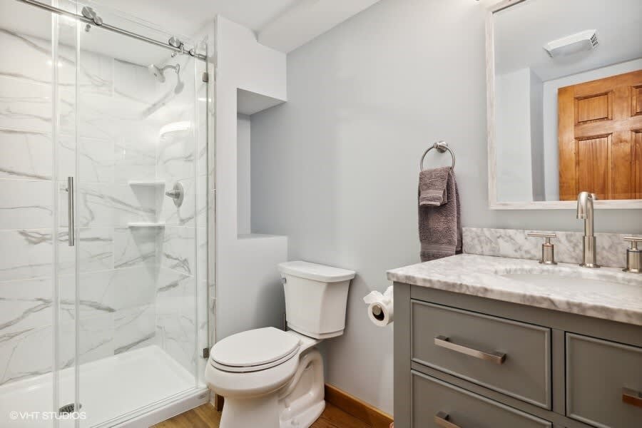 A modern bathroom features a glass-enclosed shower with white marble-patterned tiles, a white toilet, and a gray vanity with a marble countertop and a rectangular mirror. The walls are painted a light gray, and the floor has wood-look planks.