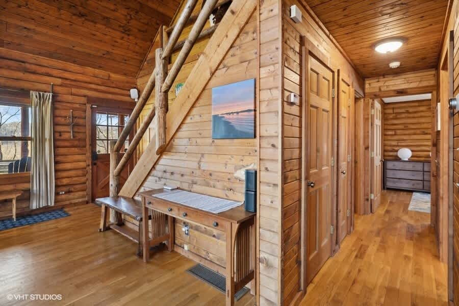 Rustic log cabin foyer with a wooden console table, a long wooden bench, and a staircase with log railings. The walls are made of horizontal wooden logs, and there are several wooden doors leading to other rooms.