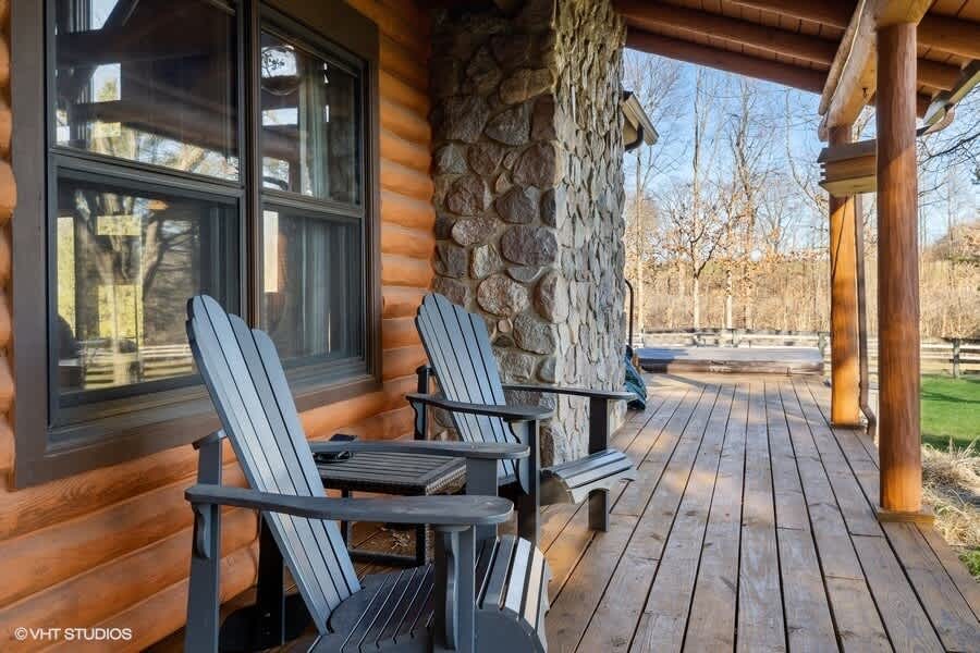 Cozy wooden porch with two dark Adirondack chairs and a small table, set against a log cabin exterior with a prominent stone column, offering a view of the surrounding trees.