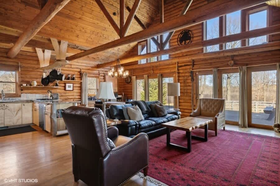 Open-concept log cabin living area with a black leather sofa, two brown armchairs, a wooden coffee table on a red patterned rug, and a kitchen with light wood cabinets and stainless steel appliances. Large windows and glass doors offer views of the outdoors.