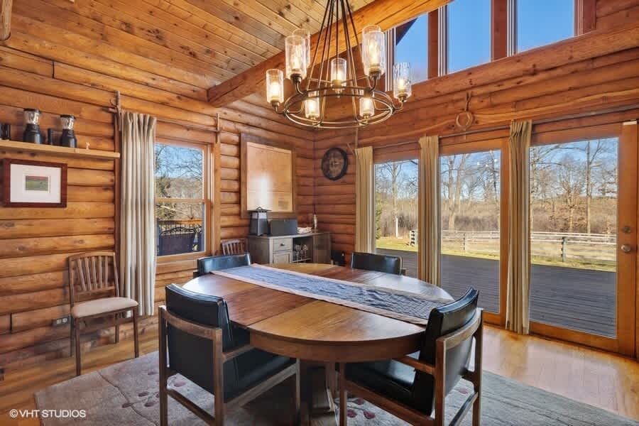 A bright dining area in a log cabin setting features a large oval wooden dining table with black leather chairs, a modern chandelier, and large glass doors opening to the exterior, with the kitchen visible in the background.