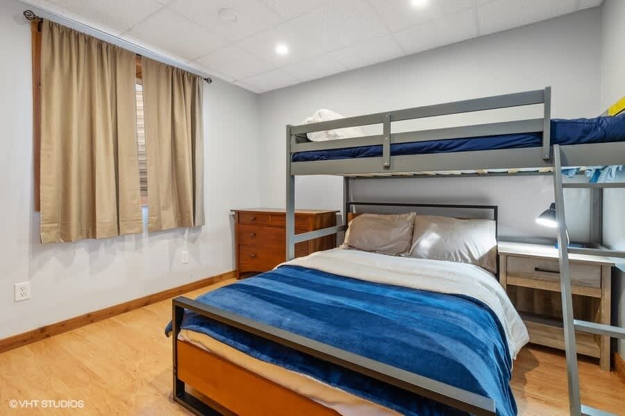 A bright bedroom features a gray bunk bed with a full-size lower bunk and a twin upper bunk, alongside a separate full-size bed with a blue striped comforter. A wooden dresser and nightstand provide storage, and a large window is covered by light brown curtains.