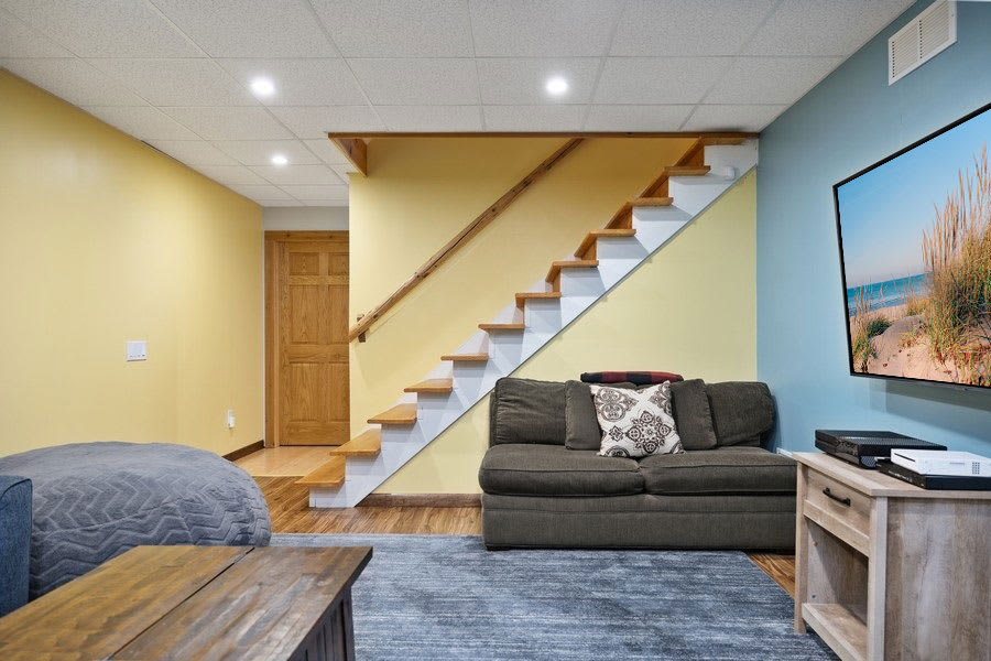Finished basement living area with light yellow and blue walls, a gray sofa, a wooden coffee table, and a large wall-mounted TV. A wooden staircase with white risers leads upstairs, and the room has recessed lighting.