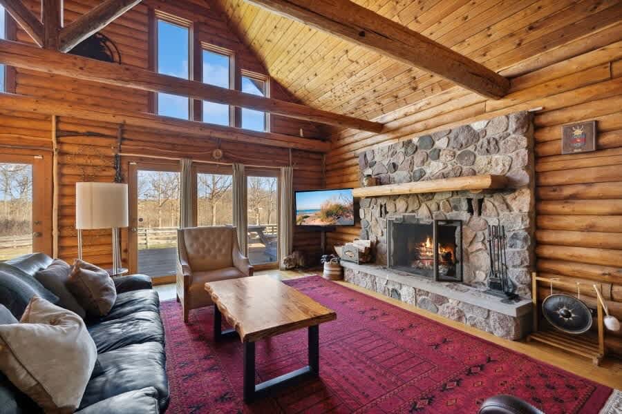 A cozy log cabin living room boasts a grand stone fireplace with a rustic wooden mantel, a dark leather sofa, a live-edge wooden coffee table, and high vaulted ceilings with exposed wooden beams.