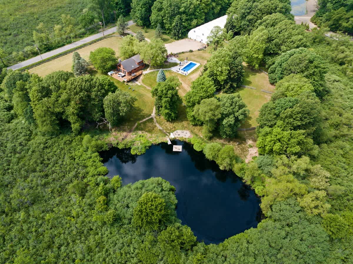 Aerial view of the property showcasing a log cabin house, a bright blue swimming pool, and a dark, natural pond with a small dock, all surrounded by lush green forest.
