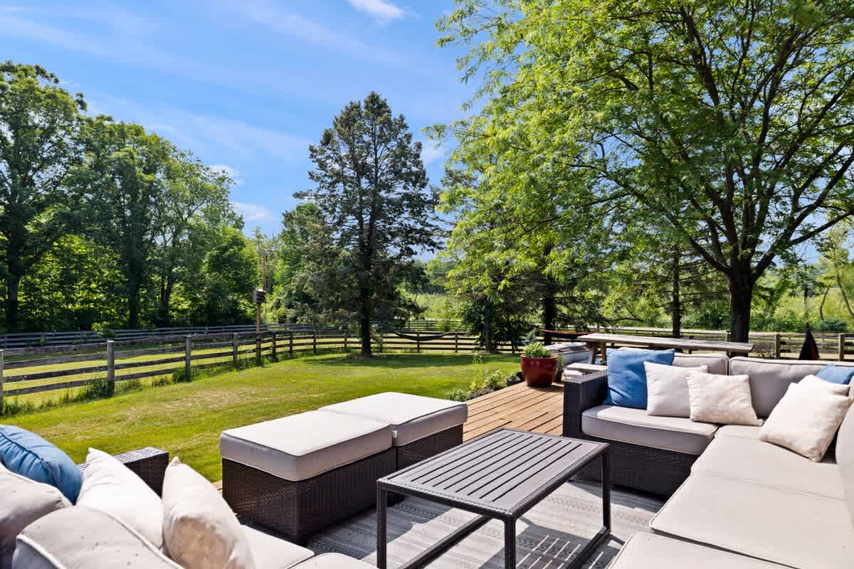 A comfortable outdoor lounge area on a wooden deck features a large L-shaped sectional sofa with light cushions and blue accent pillows, centered around a dark slatted coffee table. The deck overlooks a expansive grassy yard with mature trees and a rustic wooden fence.