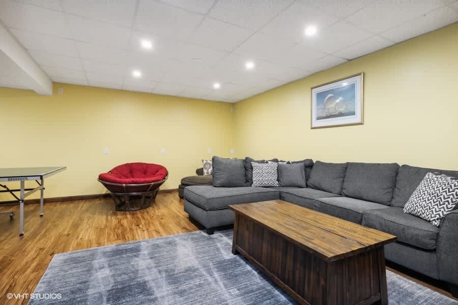 A spacious basement game room features a large gray sectional sofa, a rustic wooden coffee table, and a red papasan chair, with a ping pong table visible in the background. The room has light yellow walls, recessed lighting, and wood-look flooring.