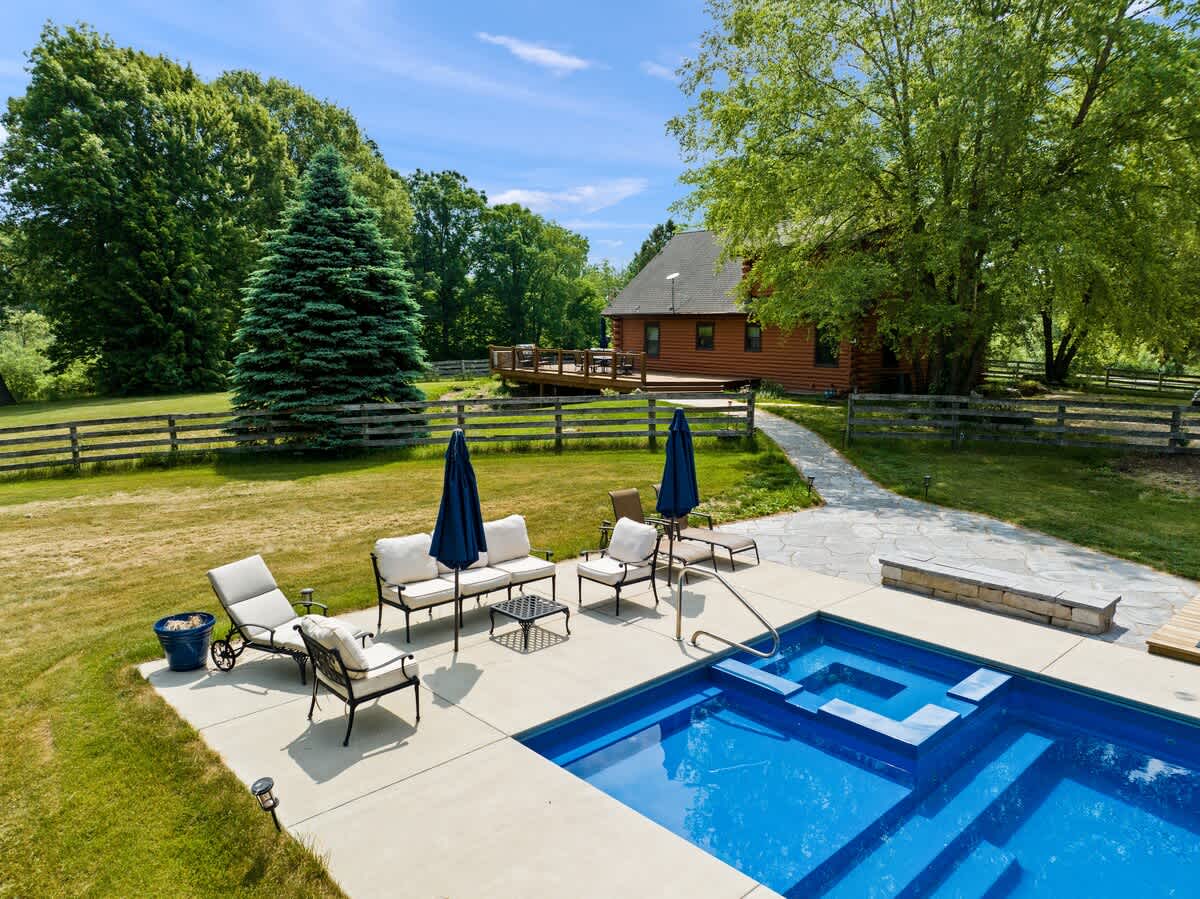 An elevated view captures a rectangular outdoor swimming pool with an integrated hot tub, surrounded by a concrete patio furnished with lounge seating and umbrellas. A stone pathway leads towards a rustic log cabin-style house with a large deck, nestled among mature trees.