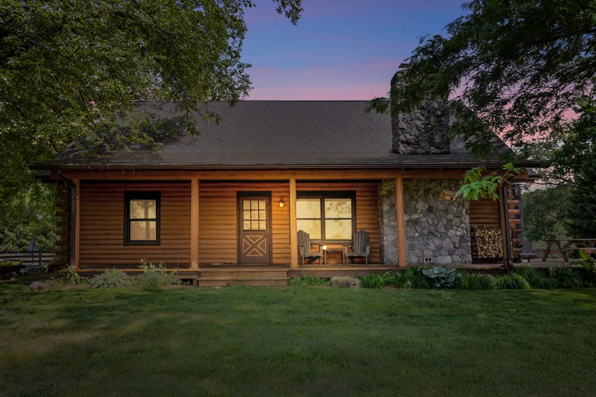 Charming log cabin house exterior at dusk, featuring a welcoming front porch with a stone chimney, surrounded by mature trees and illuminated by soft outdoor lighting.