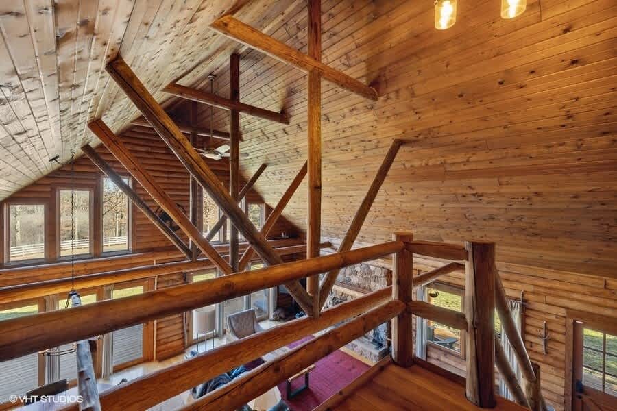 Overhead view of the log cabin living room from the second-floor landing, showcasing the vaulted wooden ceiling with exposed beams and the rustic log railings. Large windows provide ample natural light to the space below.