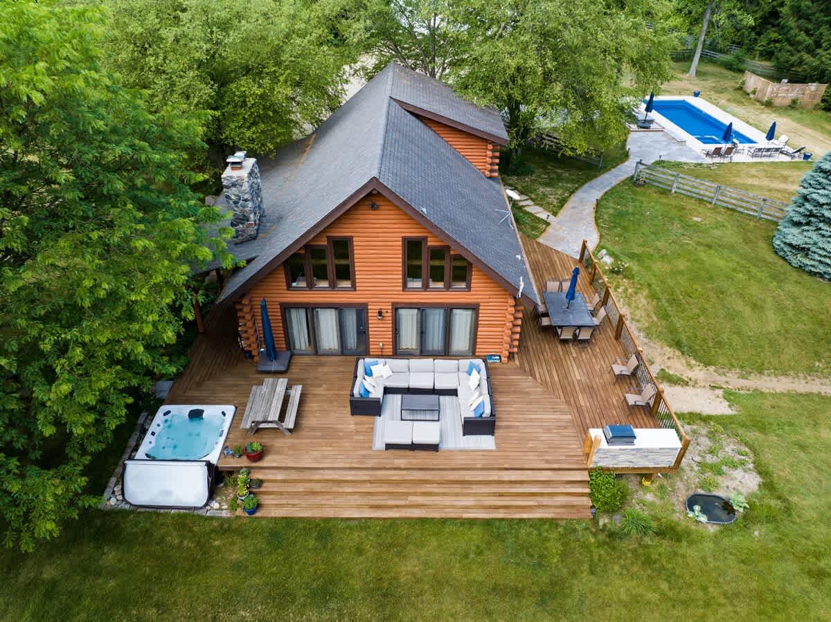 An aerial view showcases the log cabin with a large wooden deck, hot tub, and outdoor lounge area, connected by a stone path to a separate swimming pool area, all nestled among lush green trees and lawn.