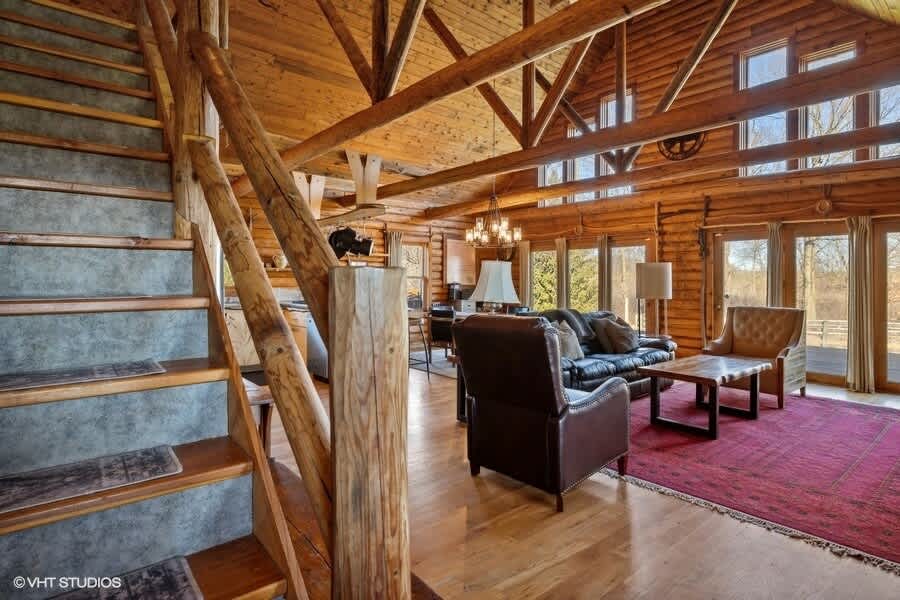View of the log cabin living room from near the entrance, featuring a rustic wooden staircase with log railings, a black leather sofa, and a red patterned rug. The room has exposed wooden beams and large windows.