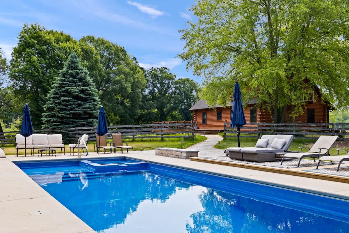 An expansive outdoor swimming pool with a shallow entry area is surrounded by a concrete deck, multiple lounge chairs, and blue umbrellas, with the log cabin and mature green trees visible in the background.