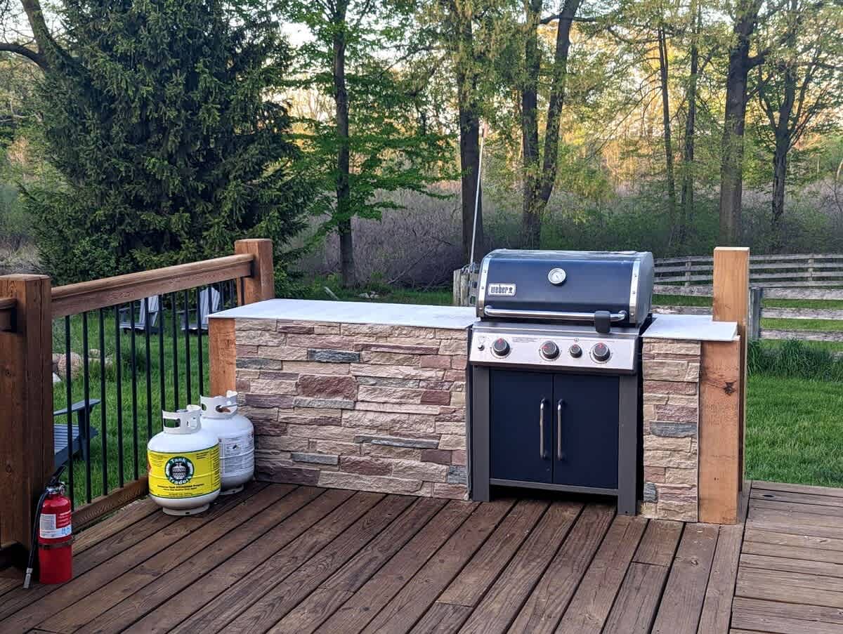 A dedicated outdoor grilling station is built into a stone-clad counter on a wooden deck, featuring a black Weber gas grill. Two propane tanks are visible next to the counter, with lush greenery and a wooden fence in the background.