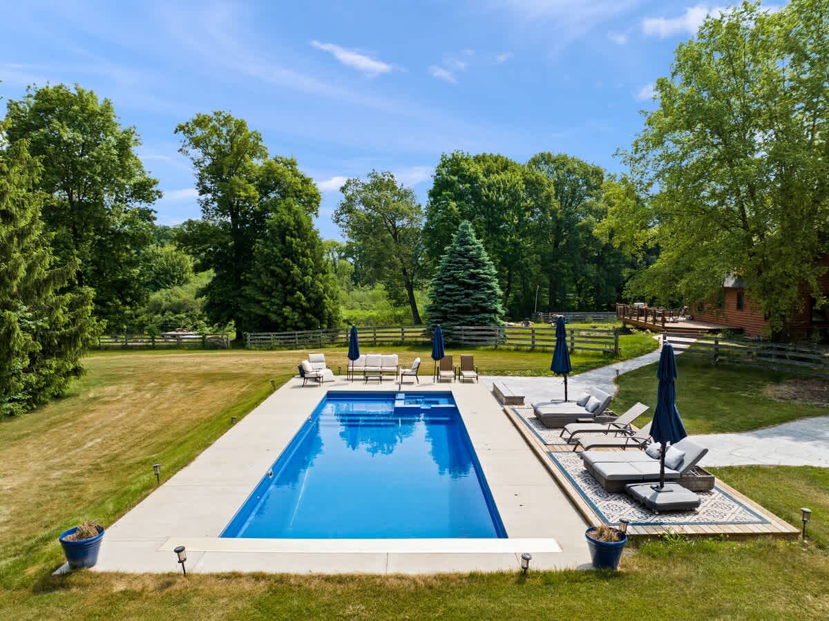 A wide aerial shot showcases a large rectangular outdoor swimming pool with a surrounding concrete patio, featuring multiple lounge chairs and blue umbrellas. The pool is situated in a vast grassy yard, framed by dense mature trees and a wooden fence, with the house visible in the distance.