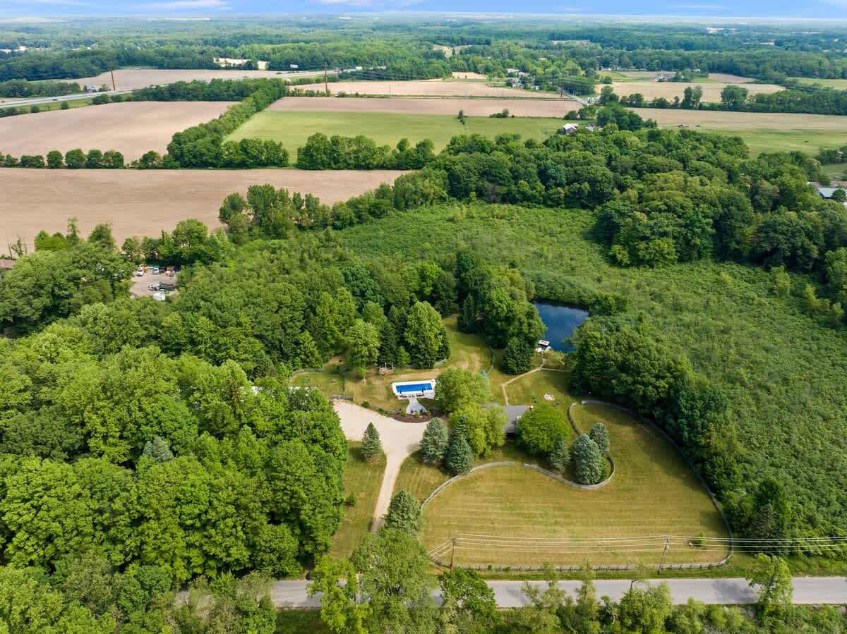 Expansive aerial view of the property, revealing a secluded house, swimming pool, and pond nestled within a large wooded area, with agricultural fields visible in the distant landscape.